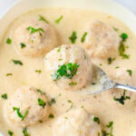 A bowl of creamy white sauce with meatballs, garnished with chopped herbs. A spoon lifts one Alfredo meatball, enticing you to savor its rich flavor.