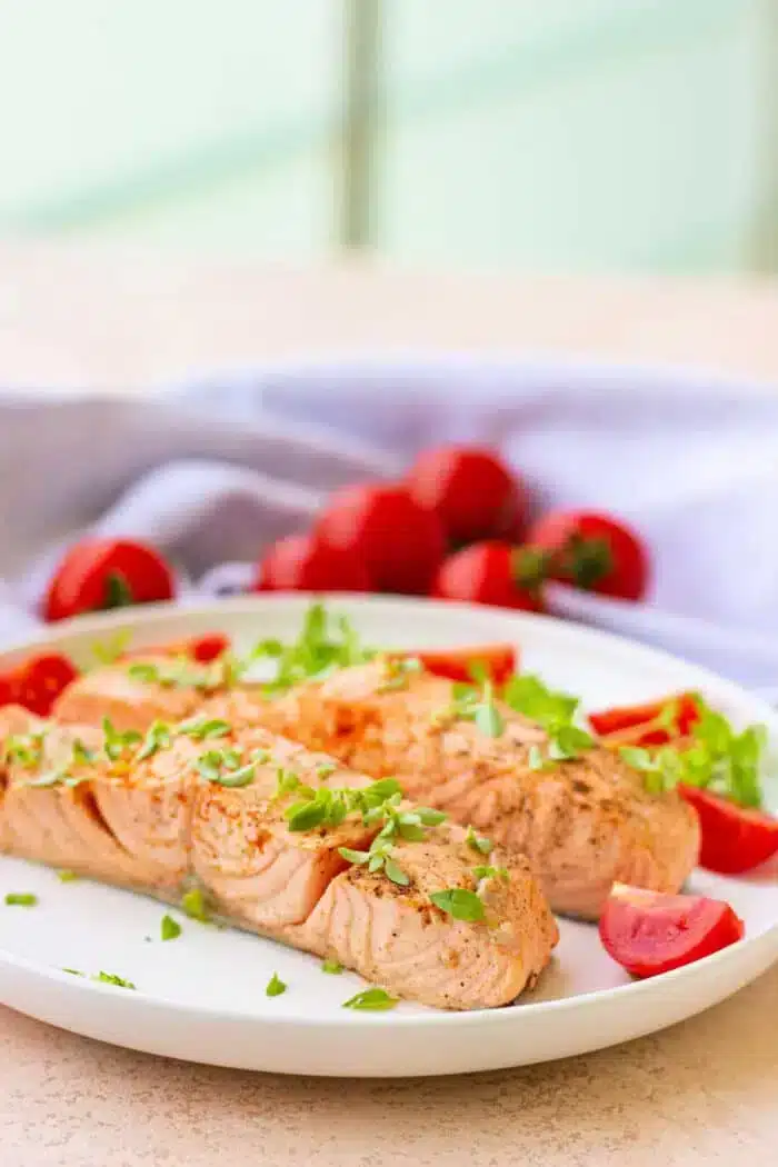 A plate with two fillets of salmon, reminiscent of delicately boiled fish, garnished with fresh herbs. Cherry tomatoes and sprigs of parsley are artfully arranged around the salmon. A light-colored cloth is in the background, along with whole tomatoes.