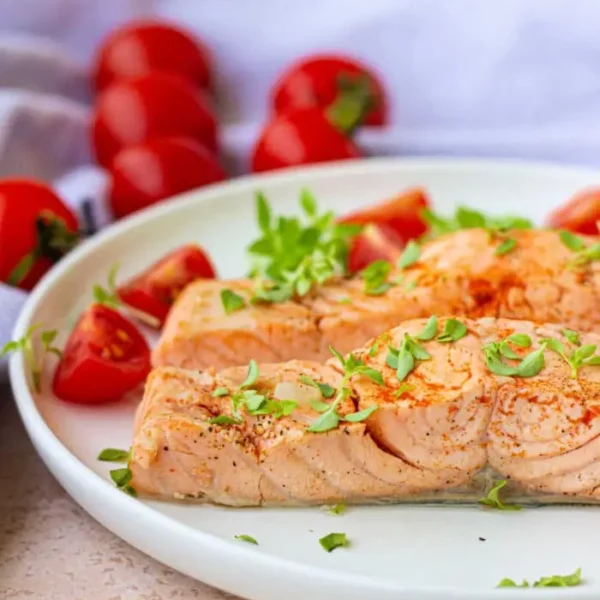 A plate with baked salmon garnished with herbs, accompanied by sliced cherry tomatoes, evokes a delightful blend of flavors. A light-colored cloth and whole cherry tomatoes sit in the background, offering a fresh contrast reminiscent of boiled fish simplicity.