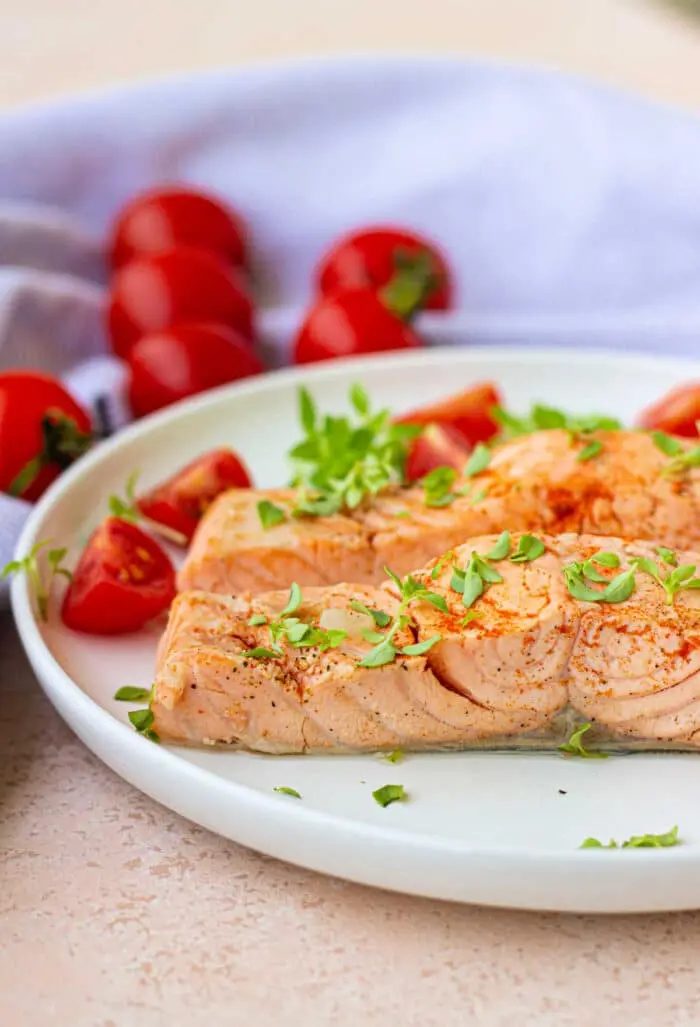 A plate with baked salmon garnished with herbs, accompanied by sliced cherry tomatoes, evokes a delightful blend of flavors. A light-colored cloth and whole cherry tomatoes sit in the background, offering a fresh contrast reminiscent of boiled fish simplicity.