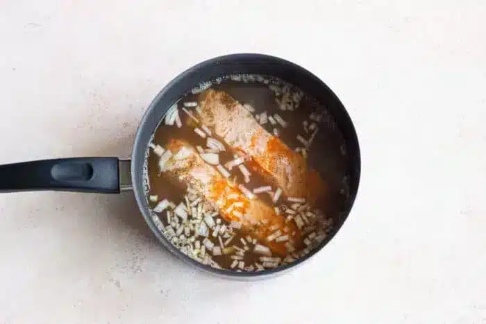 A black saucepan with fish fillets gently boiled in a seasoned broth, topped with chopped onions. The pan rests on a light-colored countertop, promising a delicious and comforting meal.