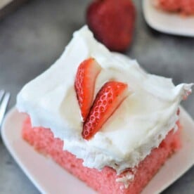 A piece of strawberry cake, made from a delicious cake mix with strawberries, is adorned with white frosting and topped with fresh strawberry slices on a plate.