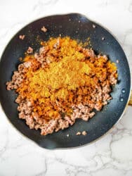 Ground meat sizzles in a frying pan, infused with the vibrant flavors of One Pot Taco Pasta seasoning, all set atop a sleek marble countertop.