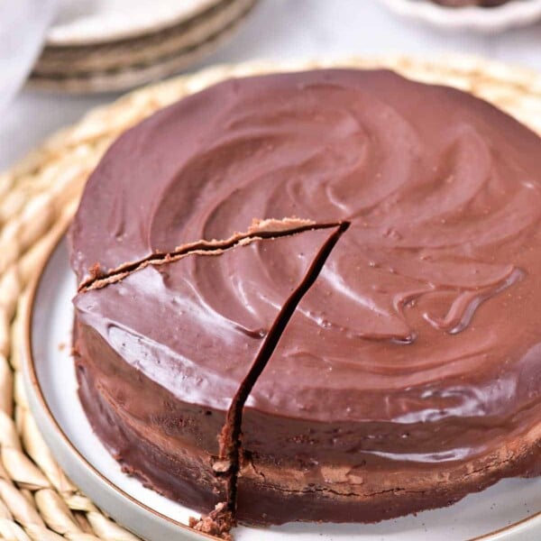 Chocolate coffee cake with a shiny ganache frosting and one slice cut out, resting on a woven placemat.