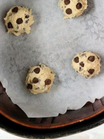 Four air fryer cookie dough balls arranged on a parchment-lined baking sheet. The dough balls are spaced apart, ready for baking. The bottom edge of a circular container is visible at the bottom of the image.