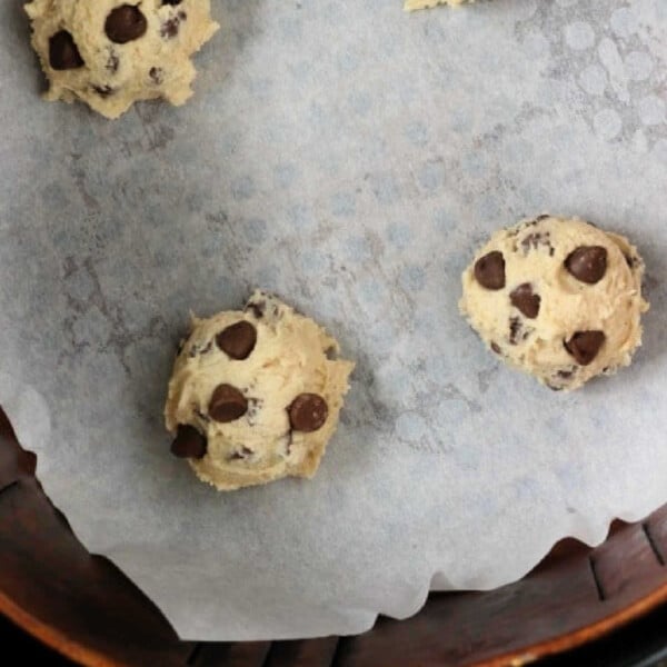 Four air fryer cookie dough balls arranged on a parchment-lined baking sheet. The dough balls are spaced apart, ready for baking. The bottom edge of a circular container is visible at the bottom of the image.