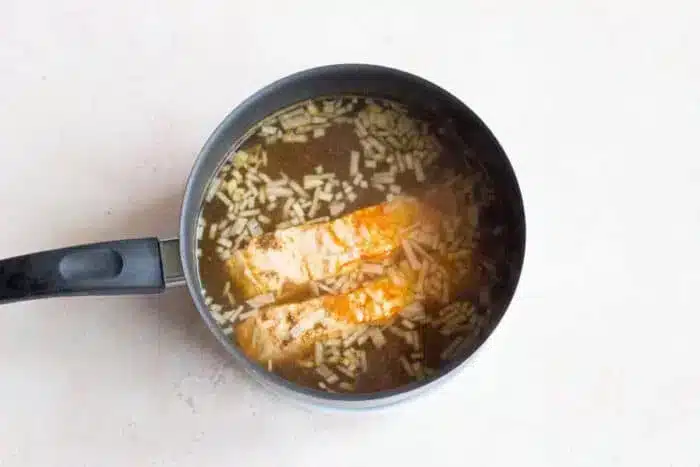 A saucepan on a light surface contains two pieces of boiled fish cooking in a broth with visible rice grains. Steam rises above the mixture, indicating it is being heated.