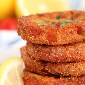 Stacked crispy fried red tomatoes are garnished with fresh herbs, served alongside lemon wedges in the background.