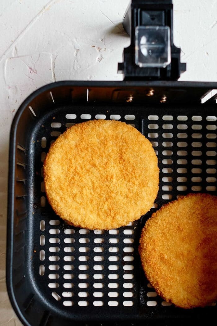 Two frozen chicken patties in an air fryer basket cook to a crispy golden brown on a light-colored surface.