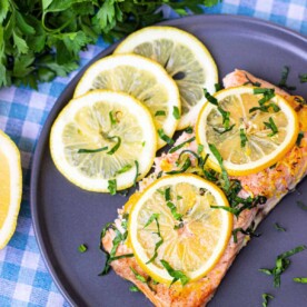 A plate of baked salmon topped with lemon slices and herbs sits on a blue and white checkered tablecloth. Fresh parsley and a sliced lemon are beside the dish, offering a hint of how long to bake salmon at 350 degrees for that perfect finish.