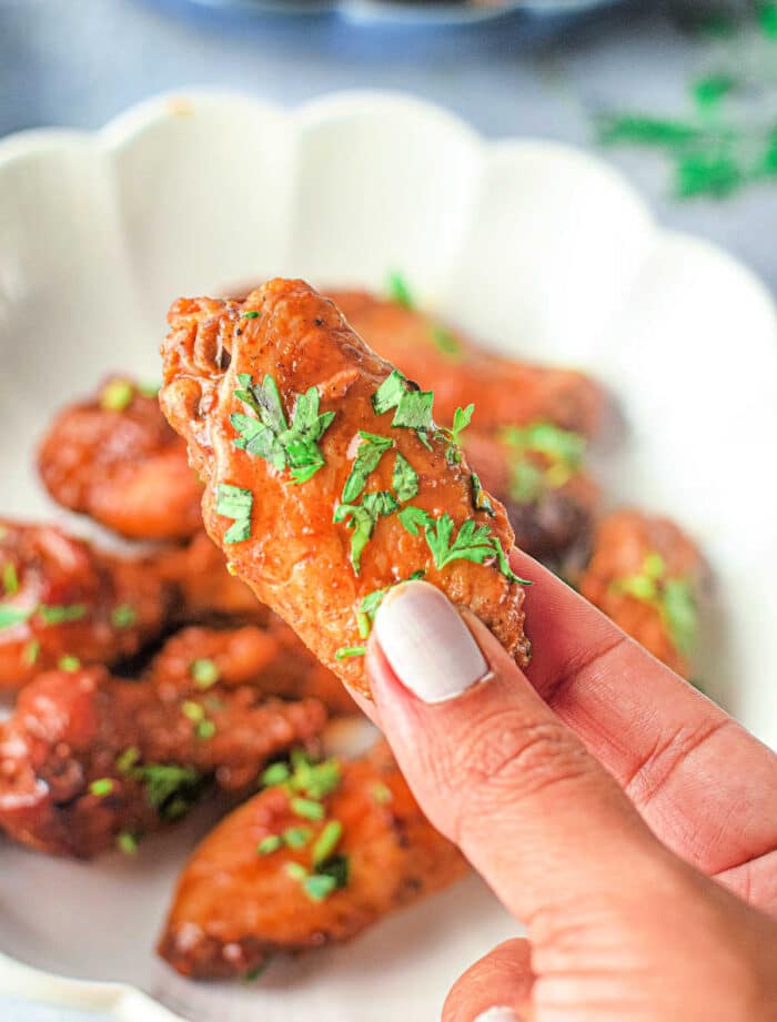 A hand holding a seasoned chicken wing garnished with fresh herbs over a white plate with additional Instant Pot Buffalo Wings in the background. The nails on the hand are painted with light-colored nail polish.