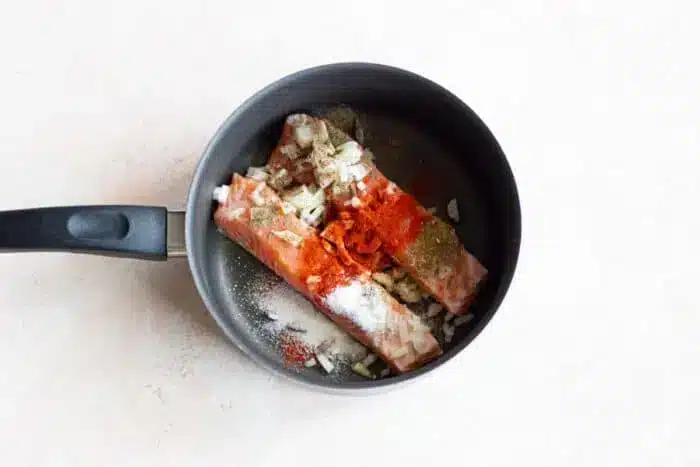 Two pieces of salmon sizzle in a pan, topped with salt, pepper, chopped onion, and red spices, capturing the essence of perfectly seasoned boiled fish.