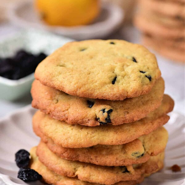 A delightful stack of lemon blueberry cookies rests on a white plate, with a fresh lemon and a vibrant bowl of blueberries adding charm to the background.