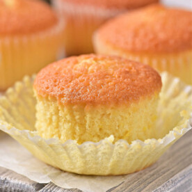 Golden vanilla cupcakes, reminiscent of a lemon muffin recipe, one with an open wrapper, rest elegantly on a wooden board.