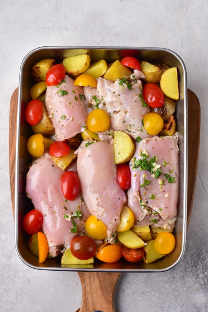 Mediterranean chicken thighs nestle among cherry tomatoes and potato slices in a roasting pan, all garnished with fragrant herbs for a delightful twist.