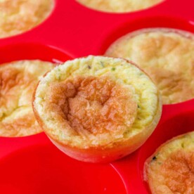 Close-up of crustless mini quiche muffins in a red silicone baking tray, one muffin slightly raised above the others.