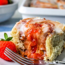 A peanut butter sweet roll with strawberry filling is topped with icing and sits on a white plate. A fork rests nearby, accompanied by a fresh strawberry. In the background, more sweet rolls can be seen in a baking dish.