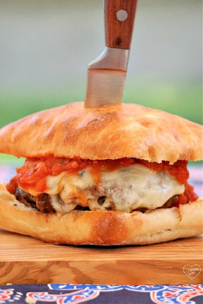 The New York-style burger, inspired by its pizza roots, features cheese and tomato sauce on a wooden board, elegantly topped with a knife.