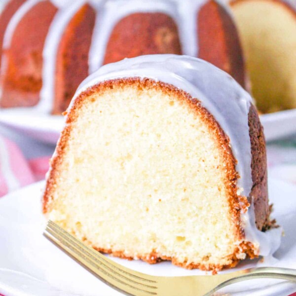 Slice of glazed bundt cake, reminiscent of a classic copycat Sara Lee Pound Cake, rests on a white plate with a gold fork, pink floral cloth underneath.