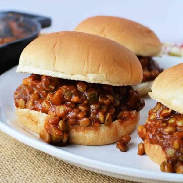 Three Sloppy Lentil Joe sliders rest on a white plate, with a skillet prominently featured in the background.