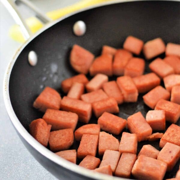 Cubed spam sizzles in a black skillet with a can of Spam in the background, perfect for starting your Spam and Eggs recipe.