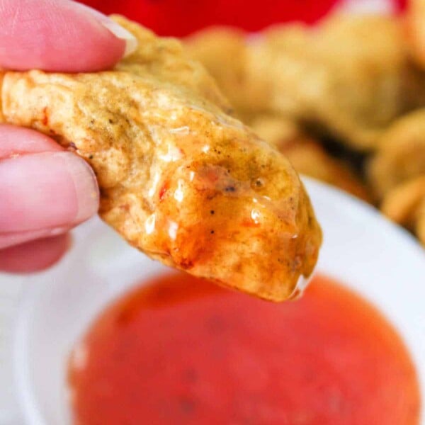 Close-up of a hand holding a golden-brown tempura chicken nuggets, dipped in a bowl of red sauce. A red cloth adds warmth to the background.