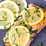 A plate with two pieces of baked salmon garnished with lemon slices and chopped herbs sits on a checkered blue and white tablecloth. The text "How Long to Bake Salmon at 350" is displayed at the top of the image, providing a useful tip.