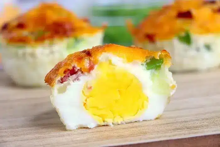 A halved egg muffin is shown on a wooden surface. The muffin has a yellow yolk center surrounded by egg white, with bits of green bell pepper, red bell pepper, and melted cheese incorporated into it, reminiscent of air fryer hard boiled eggs. Two more whole egg muffins are blurred in the background.
