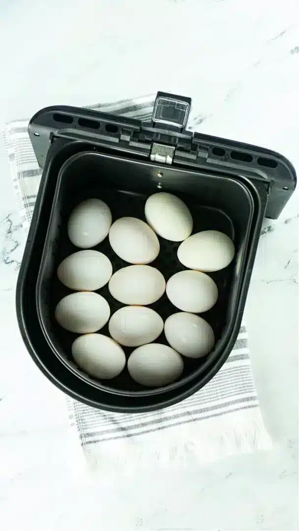 A black air fryer basket containing twelve white eggs, perfect for making air fryer hard boiled eggs, is placed on a white and gray striped cloth on a marble surface.