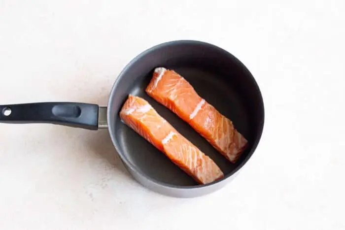 Two raw salmon fillets are nestled side by side in a non-stick pan with a black handle, ready to sizzle and infuse flavors on a light-colored surface.