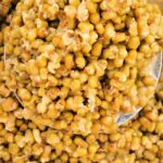 A close-up image of a spoon scooping cooked green mung beans demonstrates how to cook mung beans to a slightly mashed consistency in a thick, textured mixture. The spoon is partially visible, and the surface surrounding the beans has a rustic, slightly worn appearance.