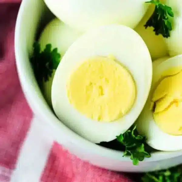 A bowl contains several air fryer hard boiled eggs, with one egg cut in half to reveal the yellow yolk. The bowl is garnished with green parsley and placed on a red and white checkered cloth.