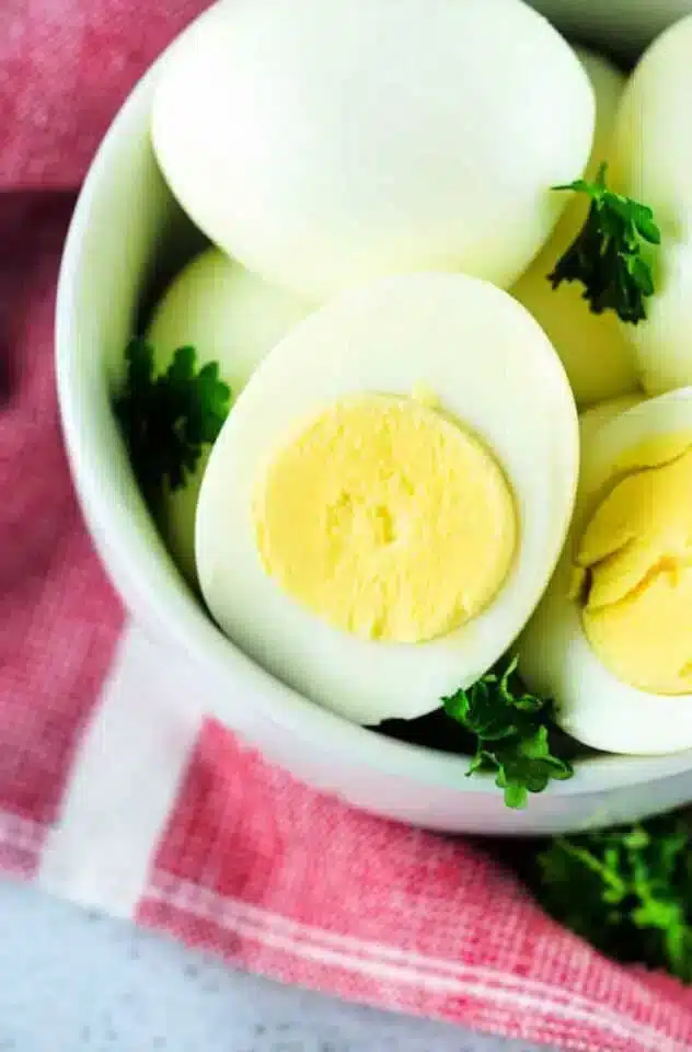 A bowl contains several air fryer hard boiled eggs, with one egg cut in half to reveal the yellow yolk. The bowl is garnished with green parsley and placed on a red and white checkered cloth.