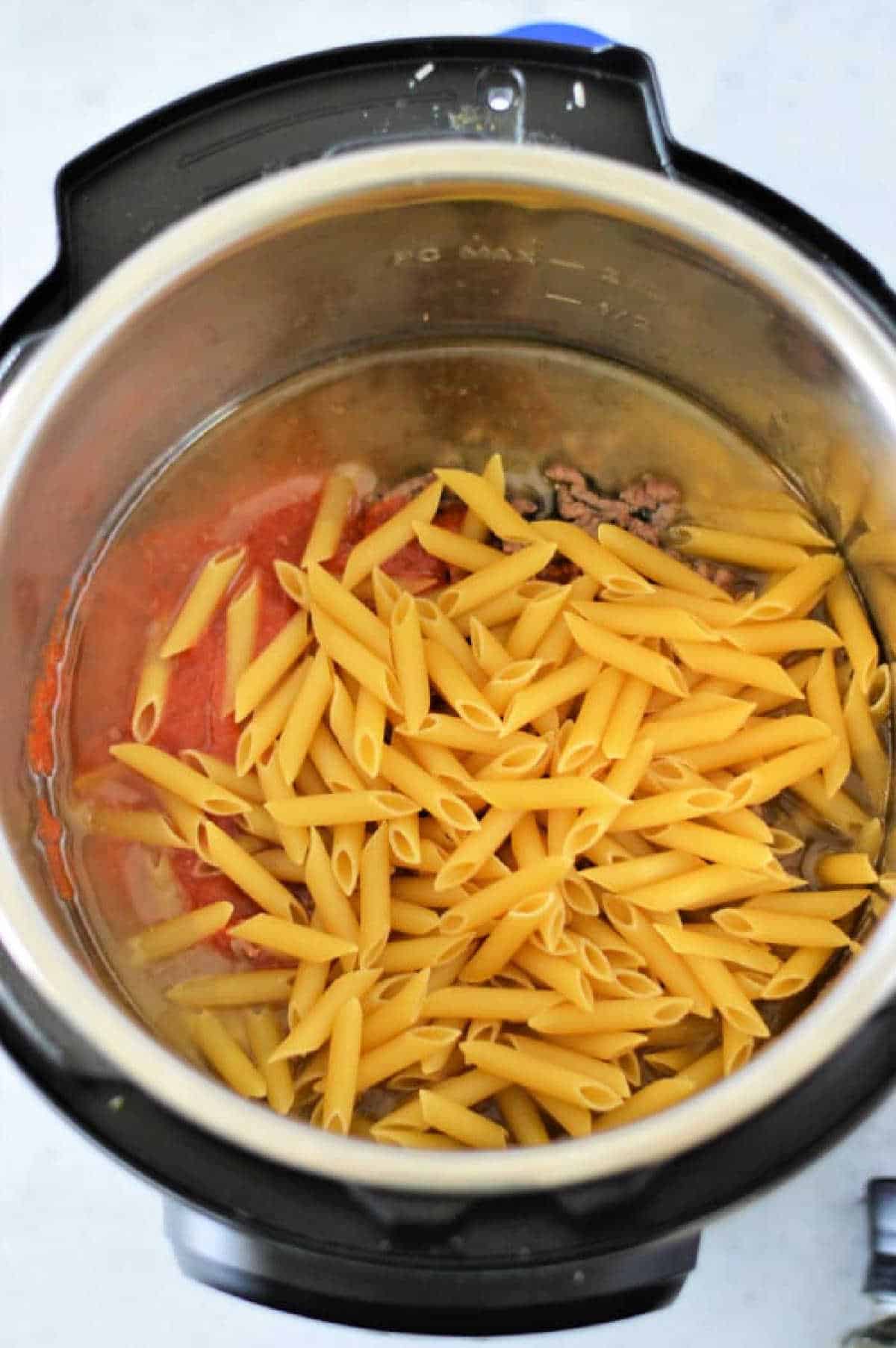 Uncooked instant pot penne pasta, ground meat, and tomato sauce are combined in the Instant Pot, ready to be cooked for a quick and delicious meal.