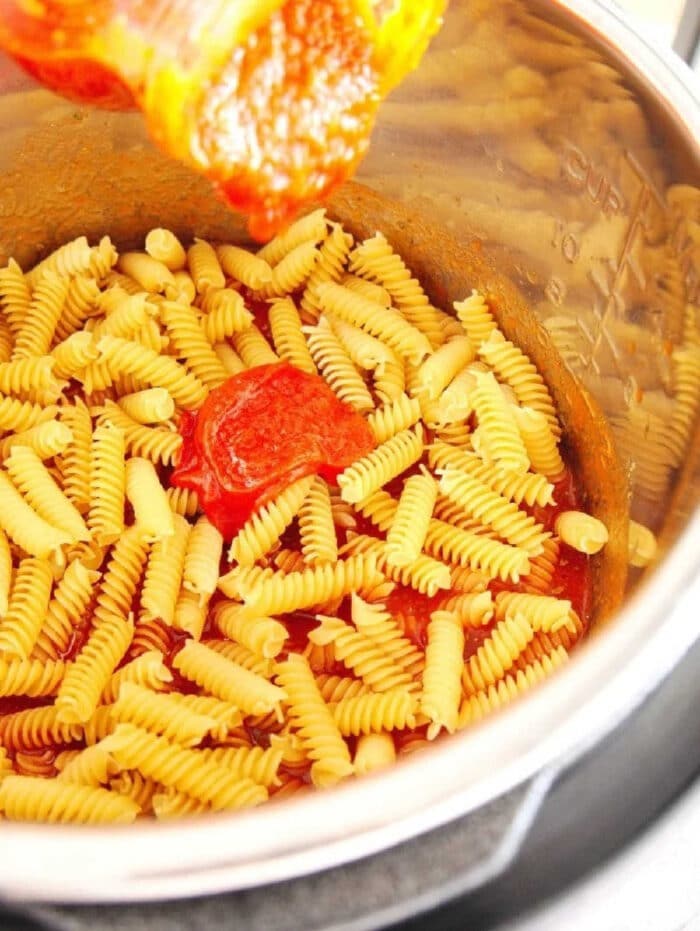 Pressure cooker pizza pasta is great! Super easy to dump it all in your pressure cooker and put in all of your favorite pizza toppings. #instantpotpizzapasta #instantpot #pizza #pasta #dinner #instantpotrecipes #thetypicalmom #pizzapasta Rotini pasta and tomato sauce in a metal pot, with sauce being poured over the uncooked pasta—perfect for making instant pot pizza pasta at home.
