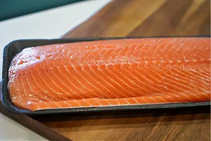 A close-up of a fresh, raw salmon fillet placed on a dark tray. The tray rests on a wooden surface, showcasing the vibrant orange color and visible marbling of what will become the best baked salmon with its rich texture and flavor.