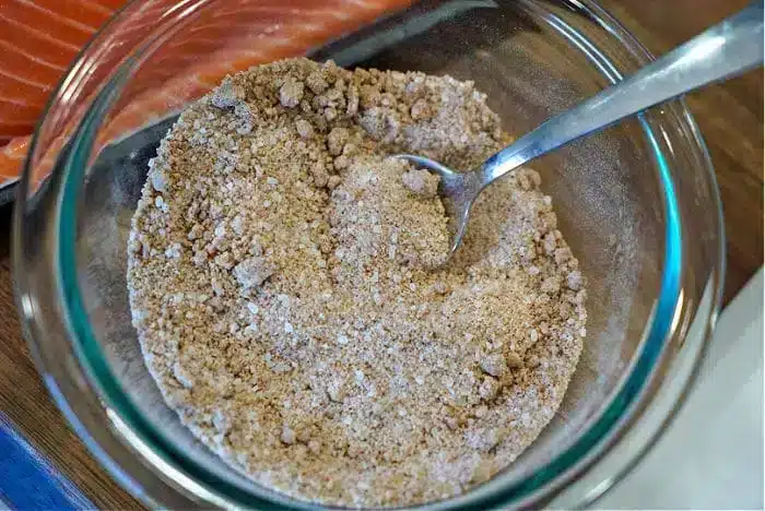 A glass bowl holds a textured, brown dry seasoning mix with a spoon nestled inside. In the background, a portion of raw salmon sits ready for transformation into the best baked salmon.