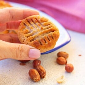 Hand holding an air fryer peanut butter cookie with a classic fork design, surrounded by peanuts on a light surface.