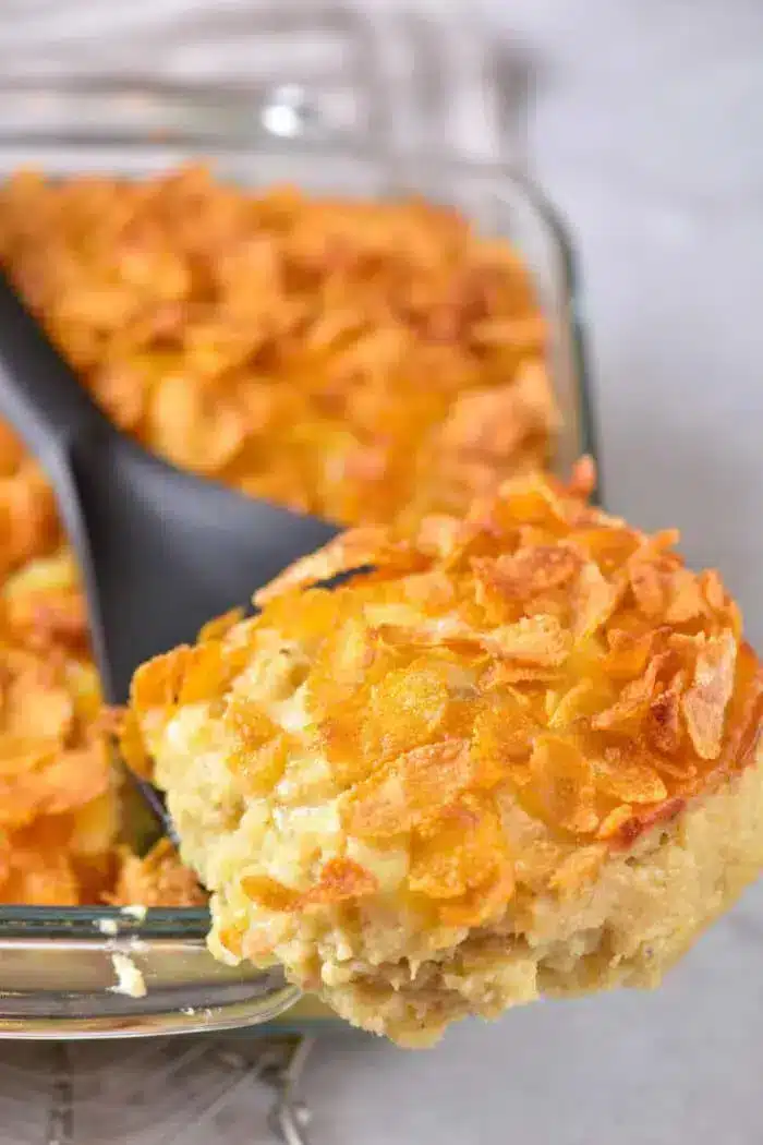 A close-up of a serving of cheesy 5 Ingredient Hash Brown Casserole being lifted from a glass baking dish with a black spoon. The casserole is topped with a layer of golden, crispy cornflakes. The background is slightly blurred.