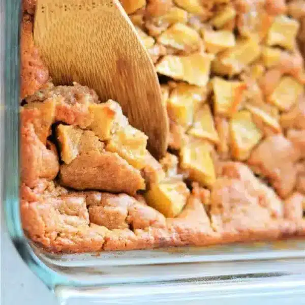 A close-up of a scrumptious Apple Cobbler with Bisquick in a glass dish. The dessert showcases tender apple pieces peeking through the golden crumble. A wooden spatula is scooping a generous portion from the corner, promising a delightful treat.