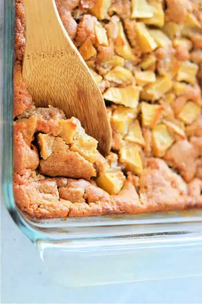 A close-up of a scrumptious Apple Cobbler with Bisquick in a glass dish. The dessert showcases tender apple pieces peeking through the golden crumble. A wooden spatula is scooping a generous portion from the corner, promising a delightful treat.