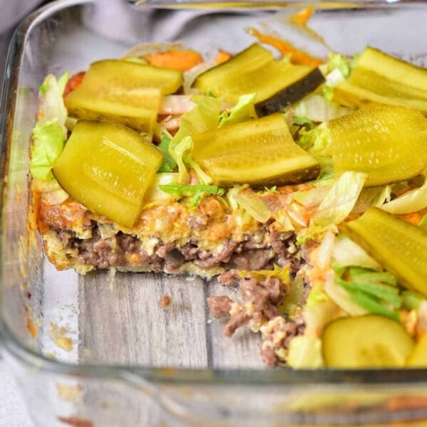 Glass dish with a layered Big Mac Casserole, topped with sliced pickles and crisp lettuce, one mouthwatering portion removed.