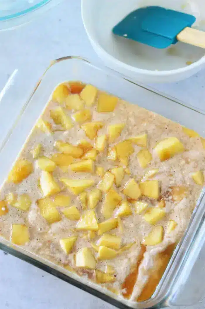 A clear square baking dish filled with a mixture of diced apples and Bisquick batter. A blue spatula rests inside a white mixing bowl in the background, ready to complete this delicious Apple Cobbler with Bisquick.