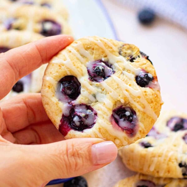 A person holding an easy biscuit with blueberries, drizzled with icing; more pastries on a plate in the background.