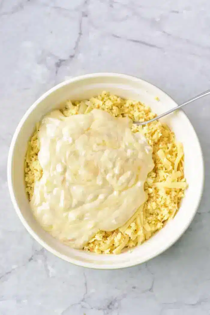 A white bowl filled with shredded cheese and breadcrumbs, topped with a thick layer of creamy sauce, conjures the inviting flavors of a 5 Ingredient Hash Brown Casserole.