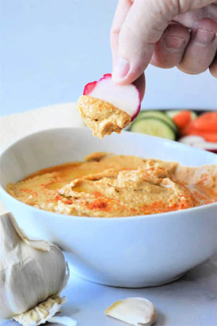 A hand dips a sliced radish into a bowl of creamy hummus made using an easy recipe with canned chickpeas, with garlic and fresh vegetables in the background.