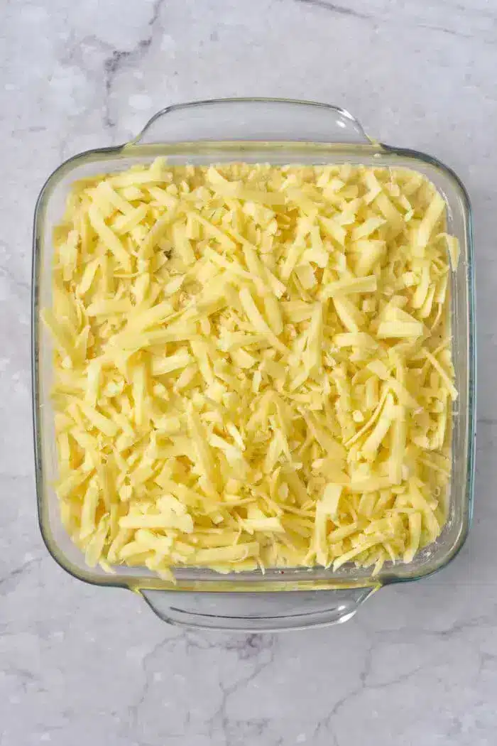 A glass baking dish filled with shredded cheese, placed on a marble countertop. The cheese is spread evenly throughout the dish.