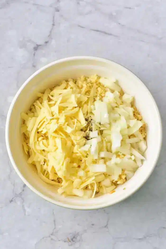 A white bowl filled with shredded cheese, chopped onions, and other finely chopped ingredients, perfect for making a delicious 5 Ingredient Hash Brown Casserole, placed on a marble surface.