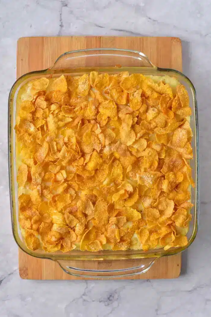 A glass baking dish filled with a 5 Ingredient Hash Brown Casserole topped with golden cornflakes sits on a wooden cutting board. The casserole has a creamy appearance beneath the crunchy cornflake topping, and the board is placed on a marble countertop.