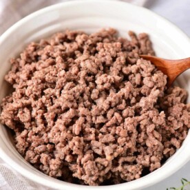 Cooked ground beef in a white bowl with a wooden spoon rests on a cloth napkin, embodying the perfect texture achieved by knowing how long to cook ground meat just right.